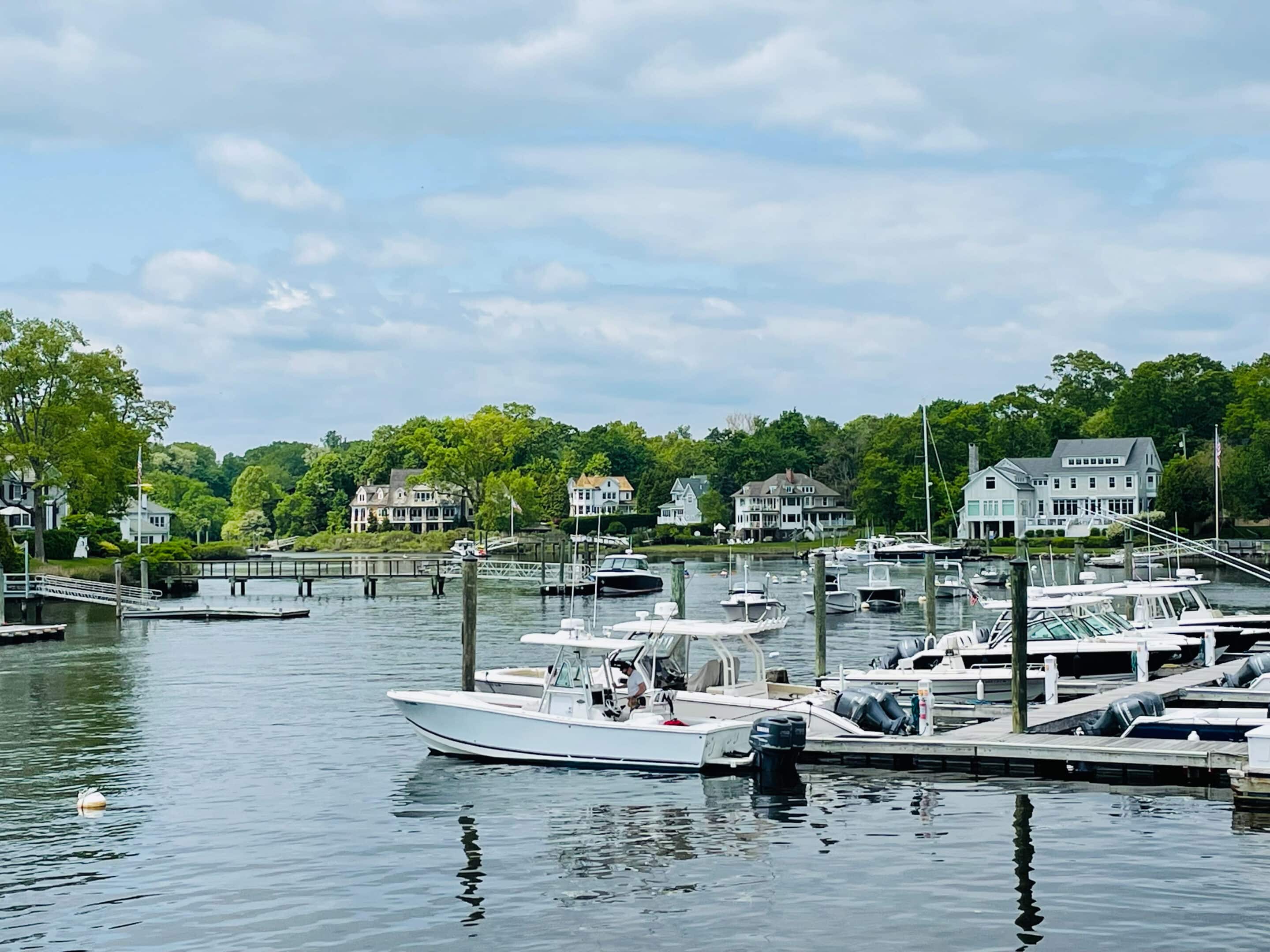 Boats docked along the waterfront in Darien, Connecticut, with coastal homes and trees in the background under a partly cloudy sky.