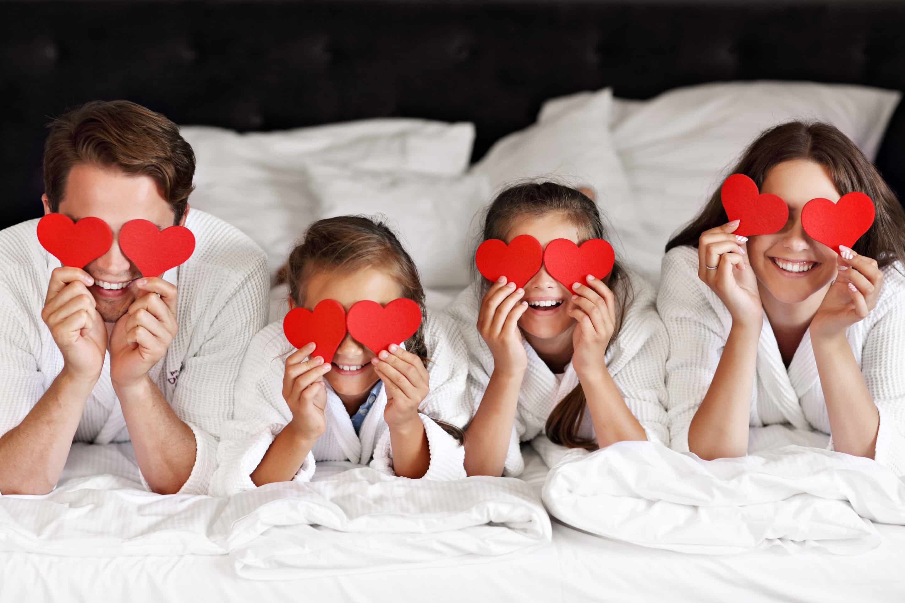 Love Where You Live – Family Valentine’s Photo Family lying on a bed holding red heart cutouts over their eyes, celebrating love and community
