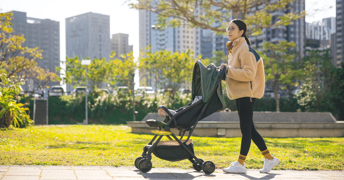 The Suburban Version of Your Favorite NYC Neighborhood Parent pushing a stroller through a sunny park with city buildings in the background, reflecting suburban-style living near NYC