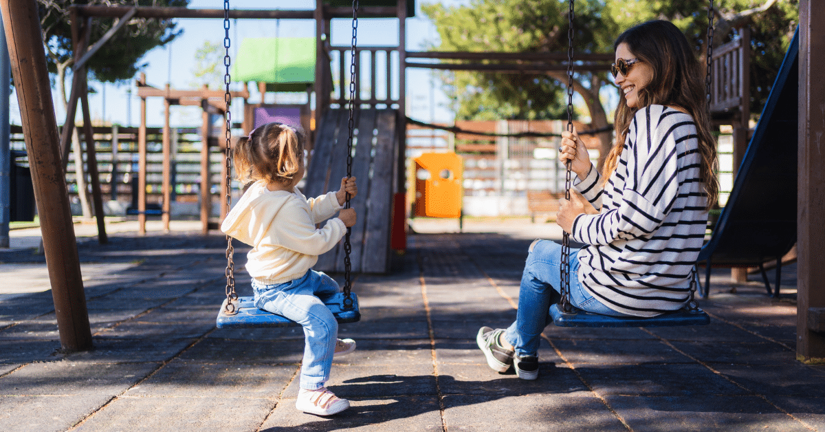 Testing a Town at the Playground A mother and young child sit on swings at a neighborhood playground, spending time together on a sunny afternoon.