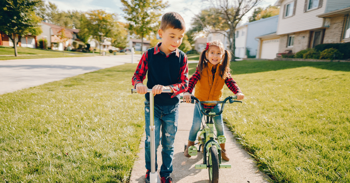 Kids Enjoying Life in the Suburbs Two children riding a scooter and bicycle along a sidewalk in a suburban neighborhood lined with homes and trees.